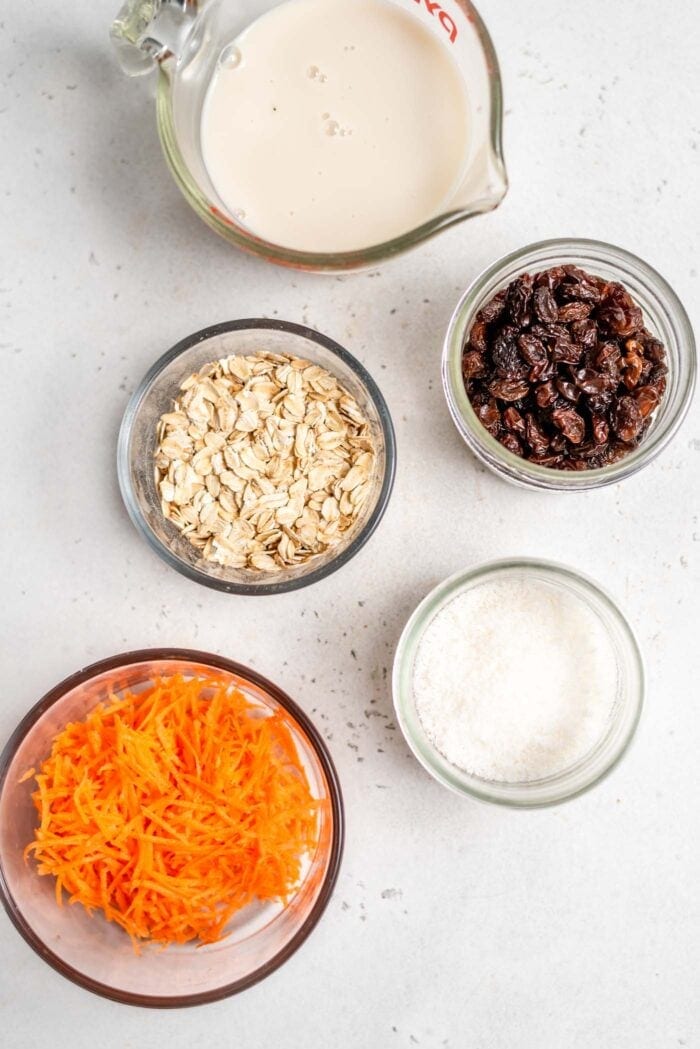 Milk, carrots, coconut, raisins and oats in containers on a white surface.