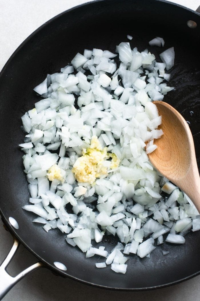 Chopped onions and garlic in a skillet with a wooden spoon for cashew cauliflower rice.