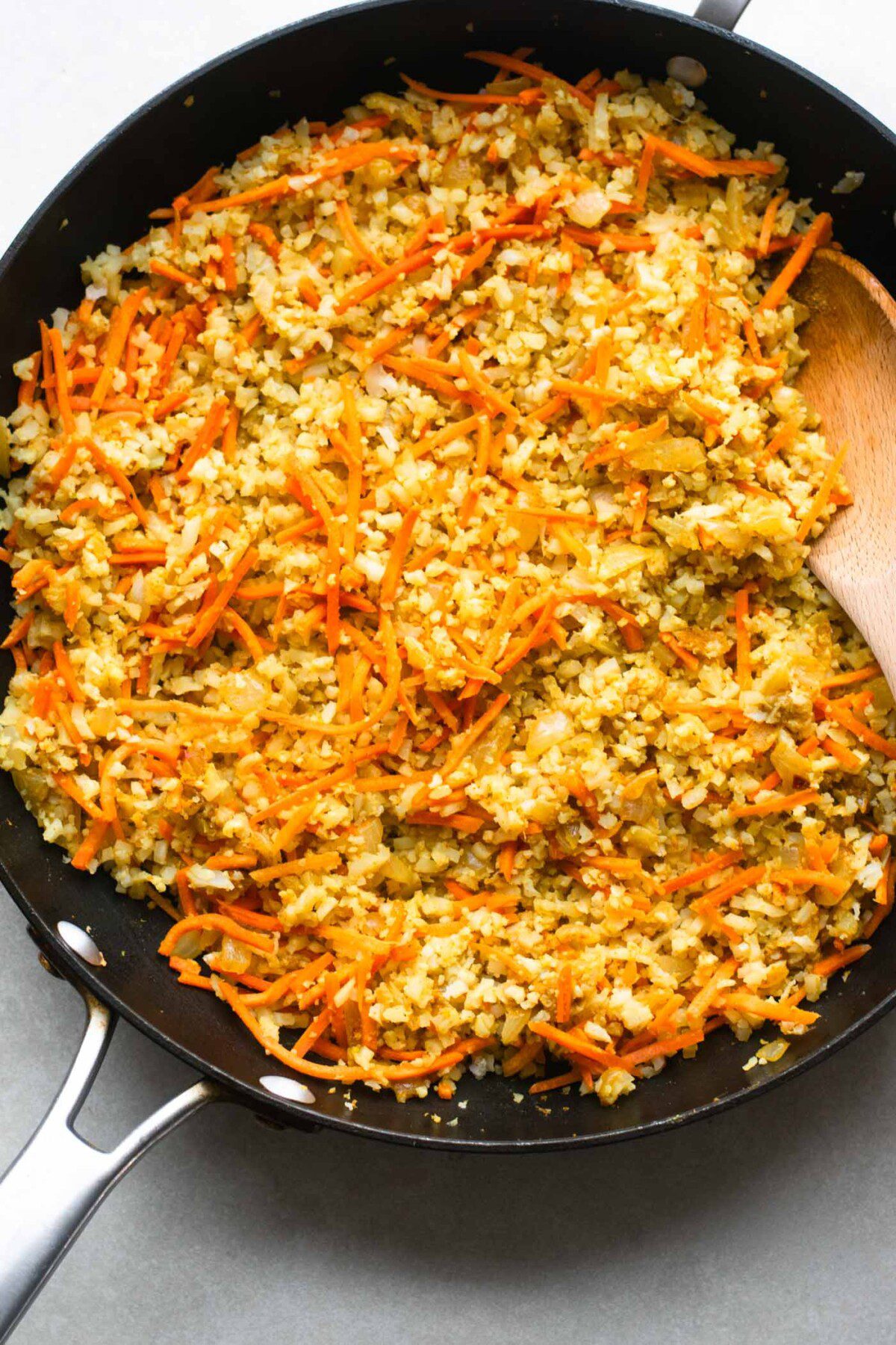 Cauliflower rice, carrots, spices, onion and garlic in a pan with a wooden spoon.