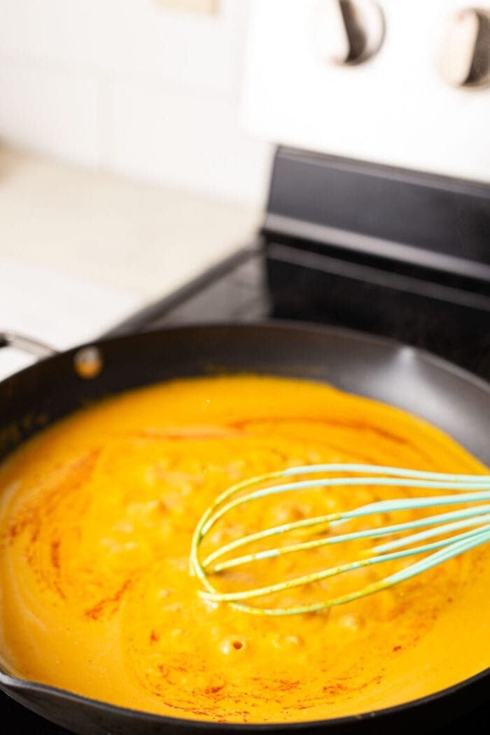 A skillet of creamy pumpkin cream sauce for pasta with a whisk resting in it on a stovetop.