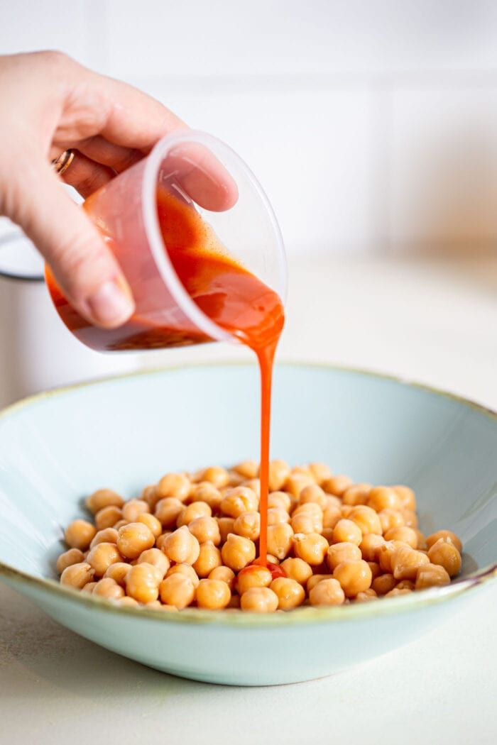 Buffalo sauce being poured into a large bowl of chickpeas.