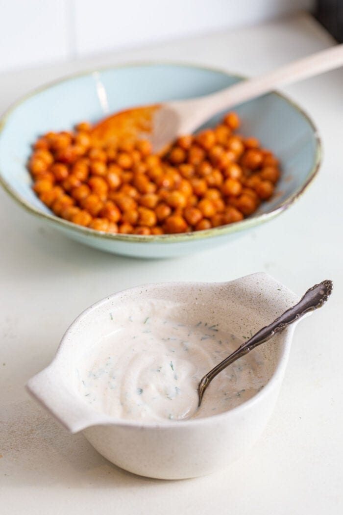 Ranch dressing in a small dish with a spoon and a large bowl of buffalo chickpeas.