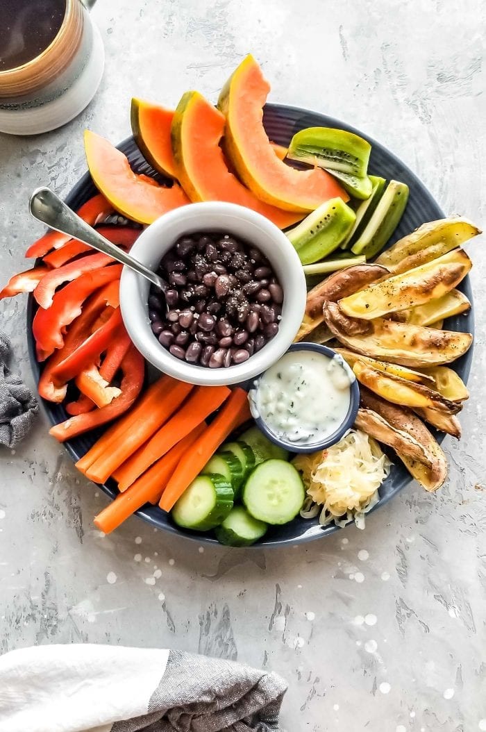 A plate with raw sliced veggies, roasted potato, black beans, papaya and a dip.