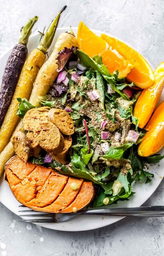 A plate with sweet potato, orange, salad, vegan sausage and roasted carrots.