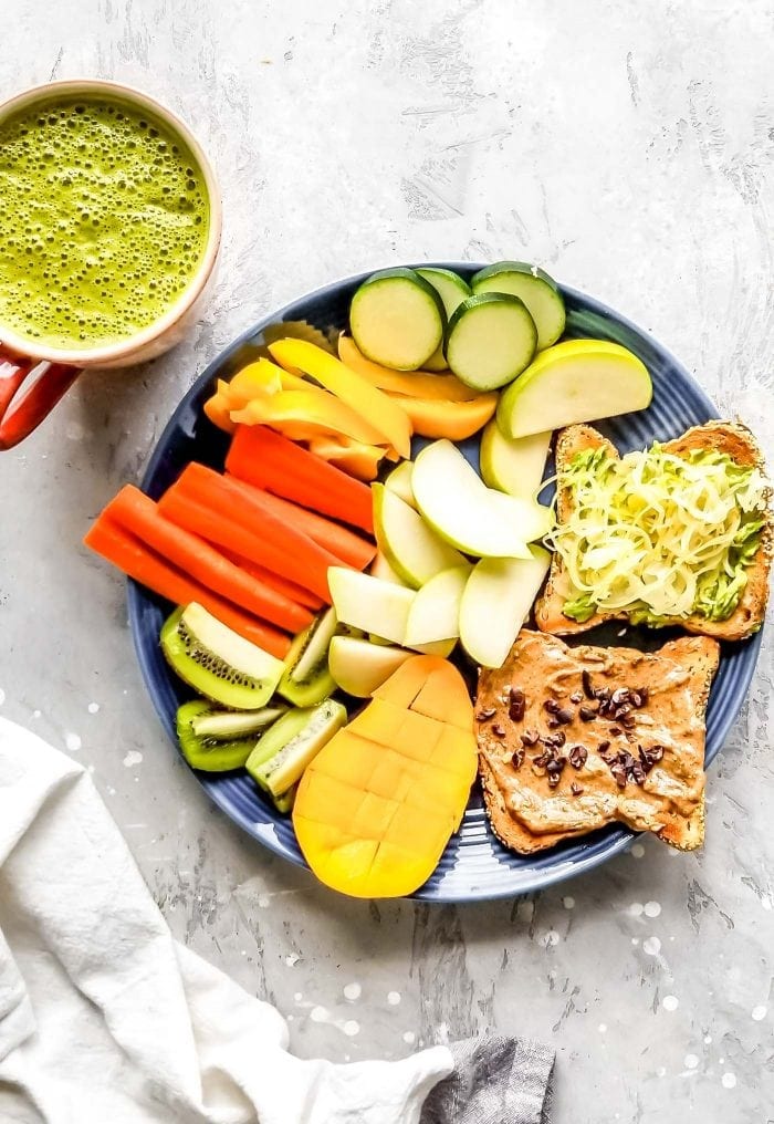 Two pieces of toast with almond butter and sauerkraut, a piece of mango, slices of kiwi and papaya, apple slices and raw zucchini slices of a plate.