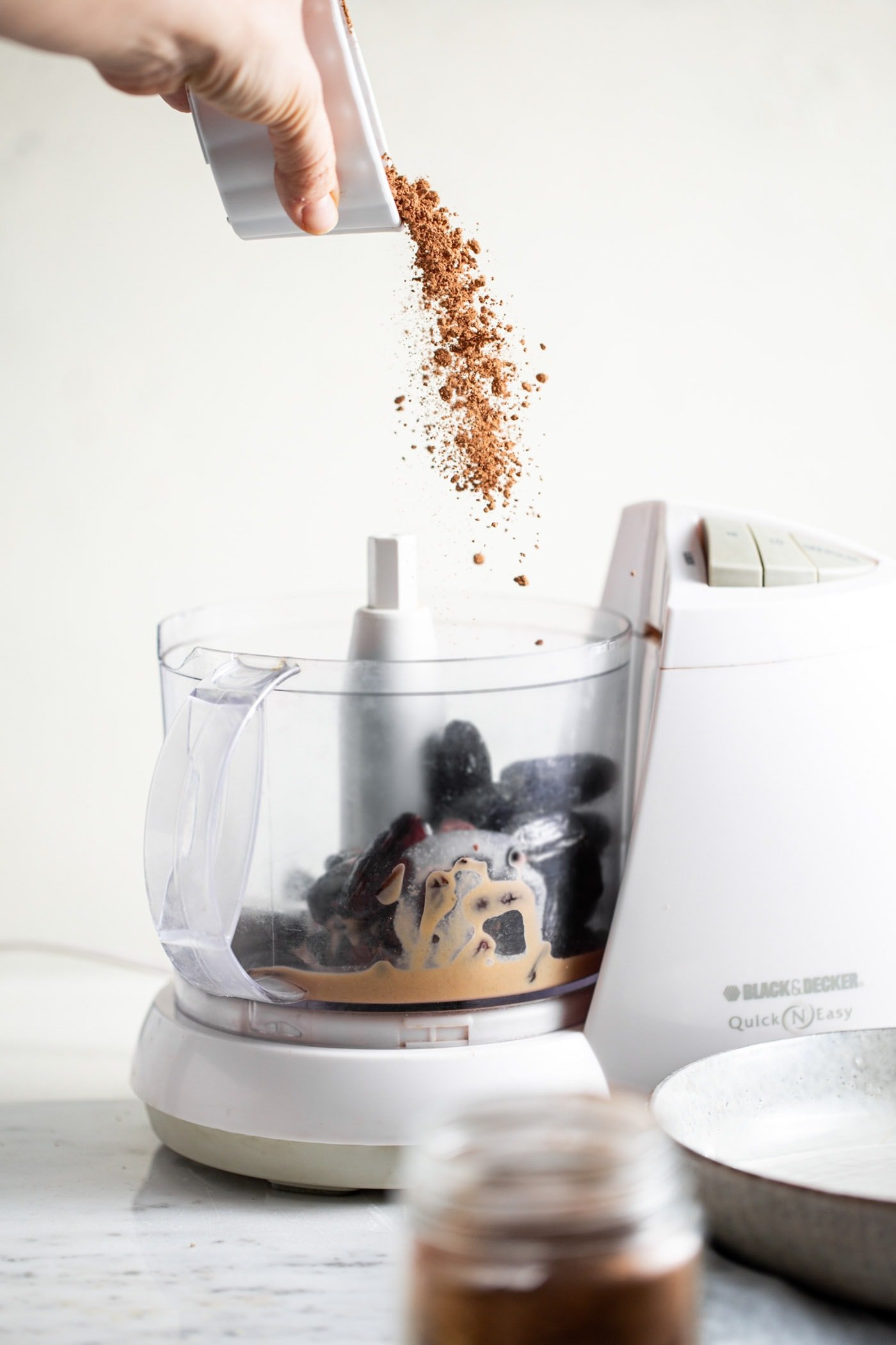 Cacao powder being dumped from a small dish into a food processor with dates and tahini.