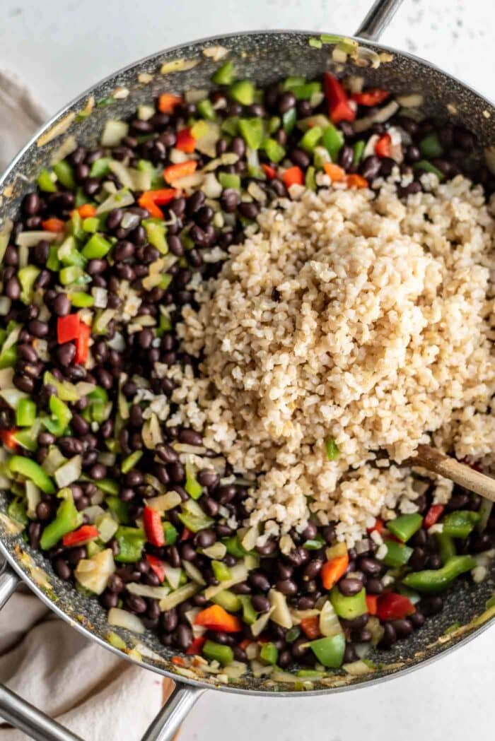A large scoop of brown rice being added to a skillet of beans and veggies.