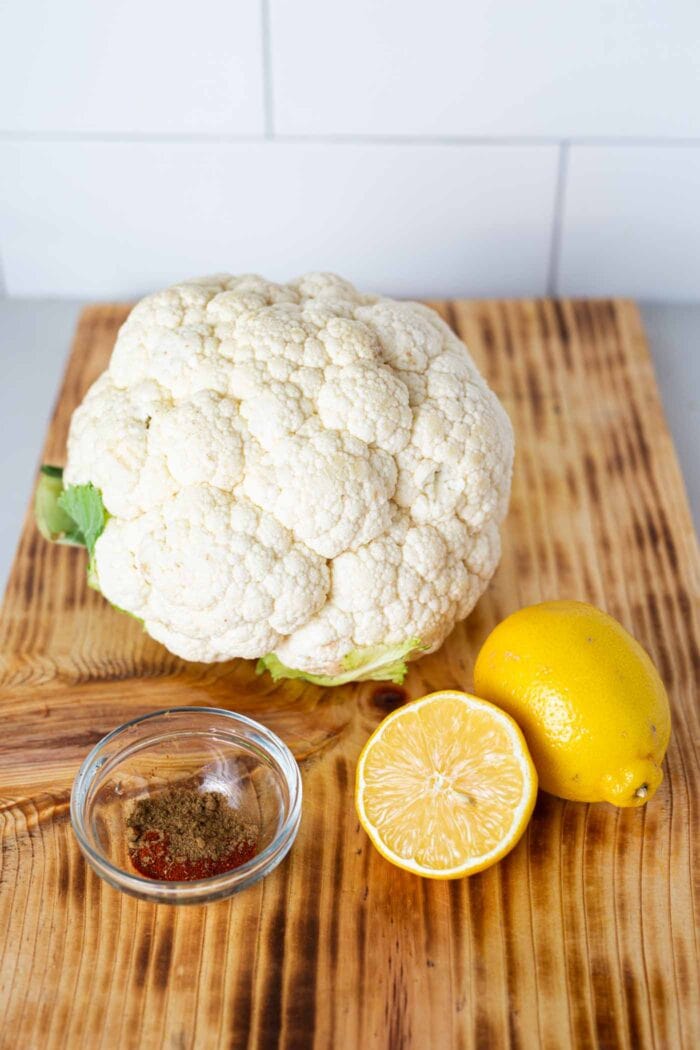 A head of cauliflower and 2 lemon wedges on a cutting board.