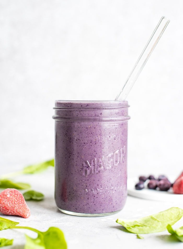 A mason jar filled with a blueberry smoothie with some spinach leaves scattered around it.