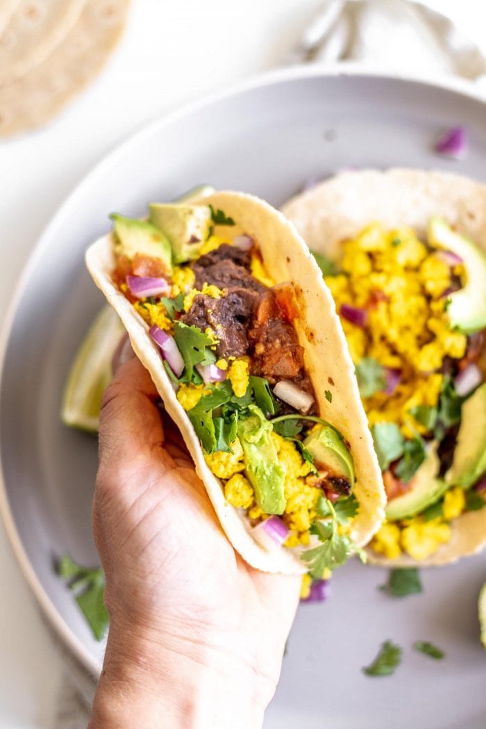A hand holding a breakfast taco with tofu scramble, refried beans, cilantro and salsa in it.