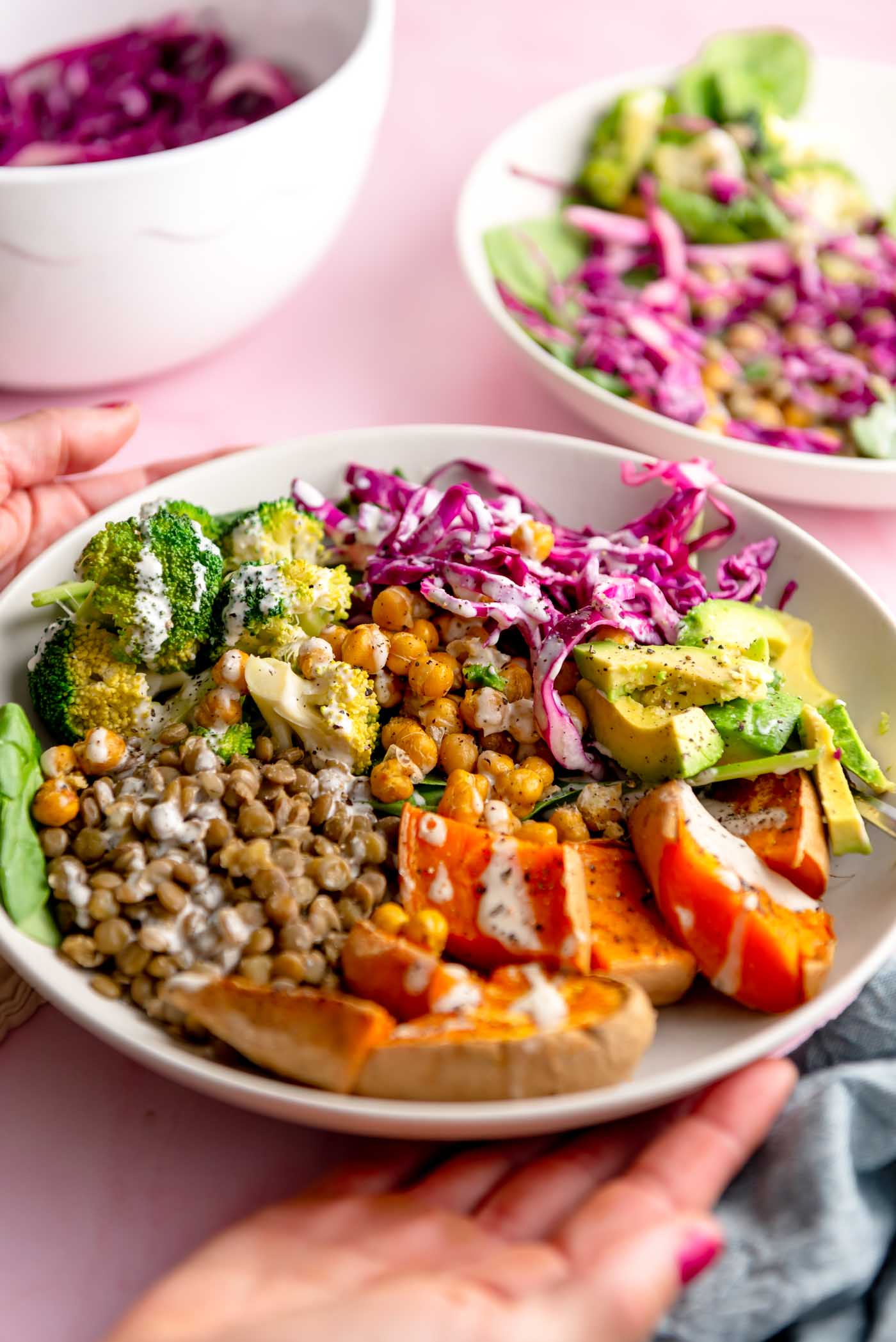 Hands holding a bowl of sweet potato, spinach, avocado, lentils, red cabbage and broccoli topped with dressing.