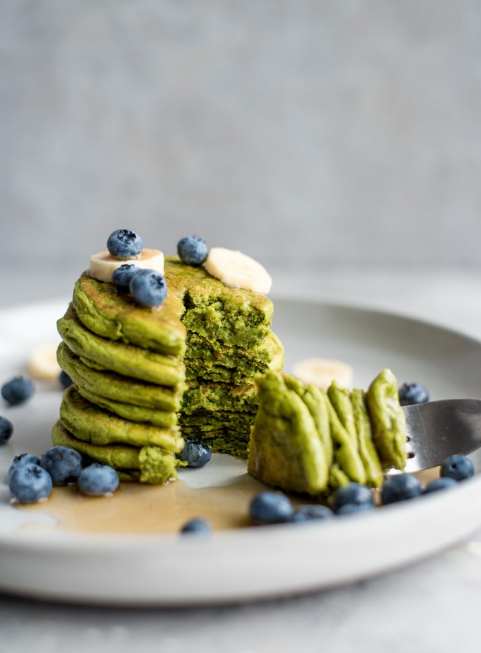 A stack of green pancakes topped with berries and banana on a plate with a fork resting on the plate.