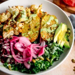 Bowl with a baked potato, chopped roasted cauliflower and chickpeas, kale, pickled red onion, shredded beets, and creamy green dressing drizzled over top.