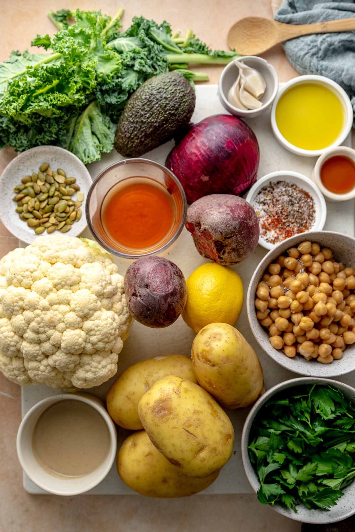 Ingredients on a cutting board to make roasted potato bowls: cauliflower, bowl of chickpeas, bowl of kale, red onion, oil, cilantro, parsley, bowl of pumpkin seeds, lemon, beets and spices.