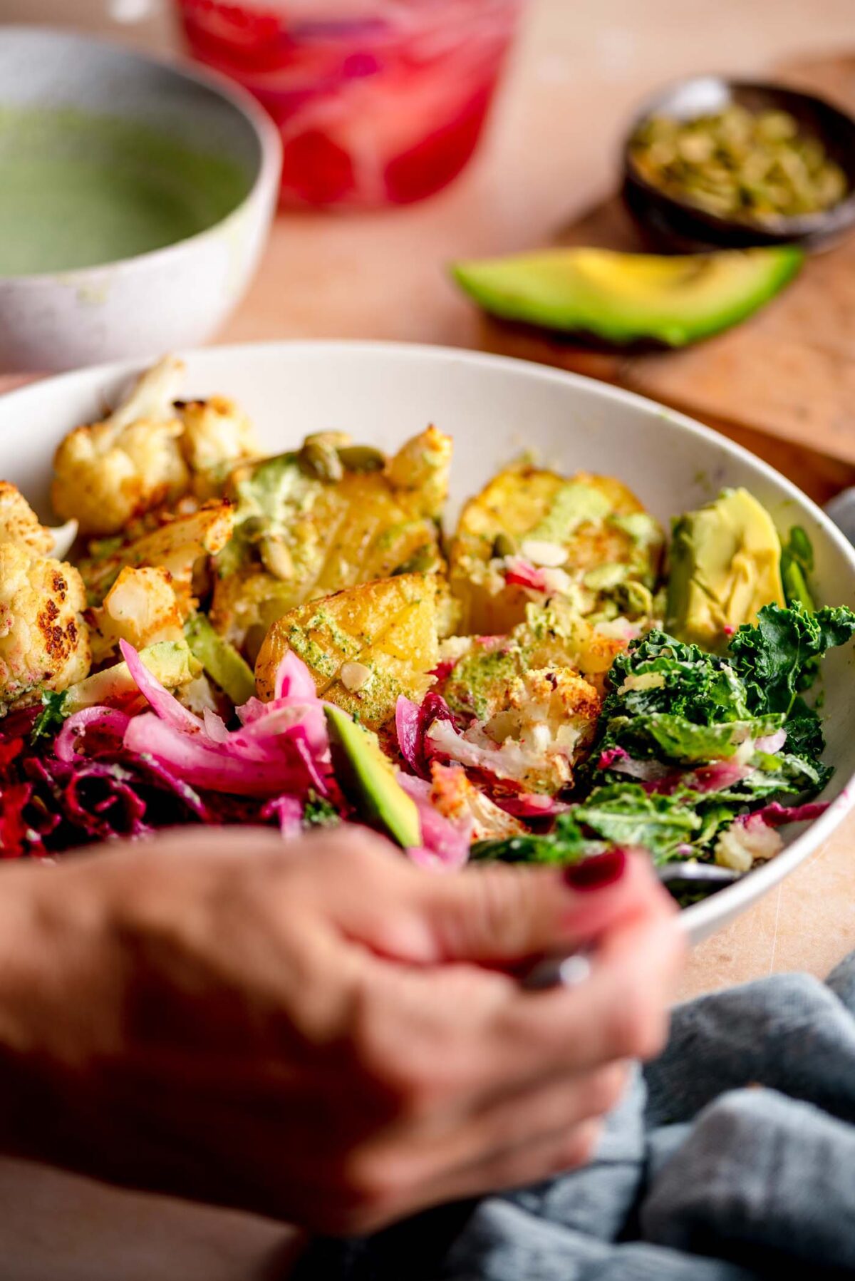 Hands using a fork in a bowl with roasted potato, pickled onion, kale, roasted cauliflower and chickpeas.