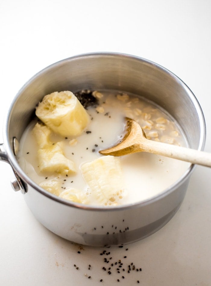 Milk, rolled oats, chia seeds and a broken up banana in a small pot with a wooden spoon.