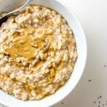Bowl of creamy oats with banana, topped with peanut butter. A spoon rests in the bowl and there are a few chia seeds scattered around.