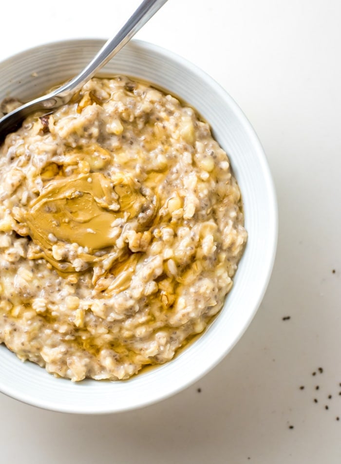 Bowl of creamy oats with banana, topped with peanut butter. A spoon rests in the bowl and there are a few chia seeds scattered around.