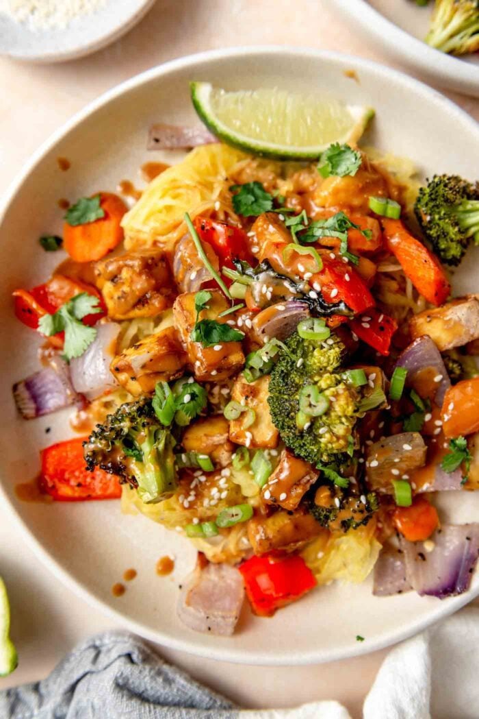 Plate of spaghetti squash noodles with peanut sauce, tofu cubes, roasted vegetables, sesame seeds and green onion.