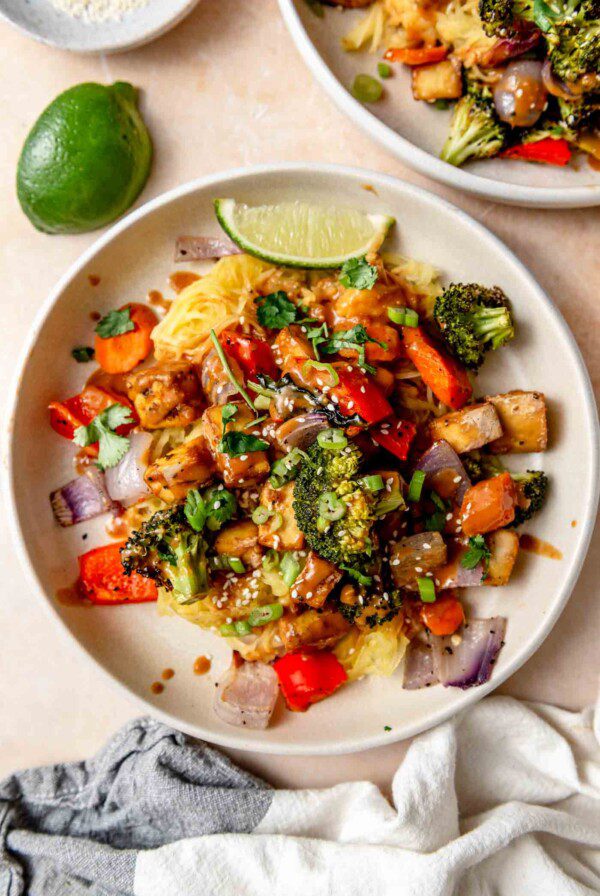 Plate of spaghetti squash noodles with peanut sauce, tofu cubes, roasted vegetables, sesame seeds and green onion.