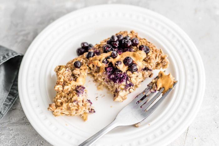 Healthy Baked Oat Recipe on a small white plate with a fork, peanut butter and fresh blueberries.