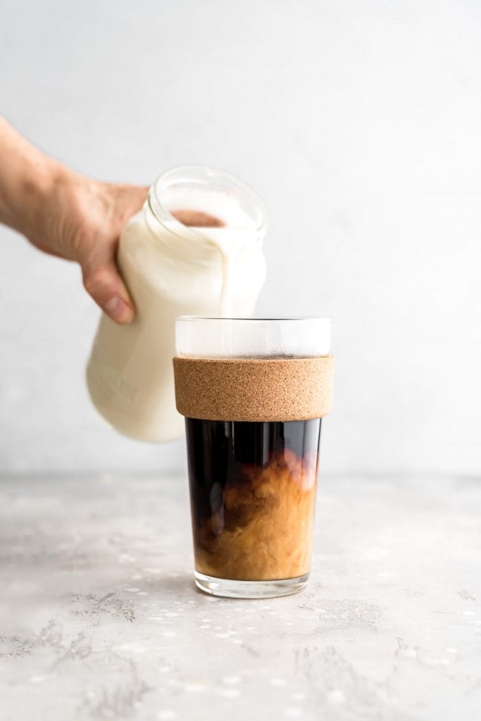 Pouring homemade oat milk into a glass of hot coffee.