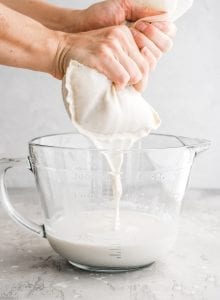 Straining the blended oat milk through a nut milk bag into a glass measuring cup.