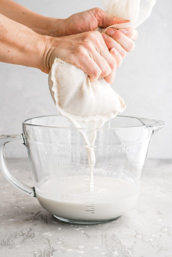 Straining the blended oat milk through a nut milk bag into a glass measuring cup.