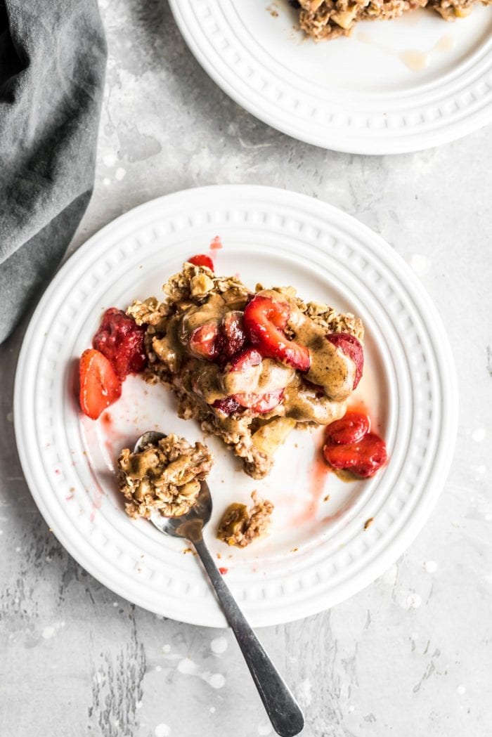 A square of baked apple oatmeal topped with almond butter and strawberries on a plate with a fork resting on it.