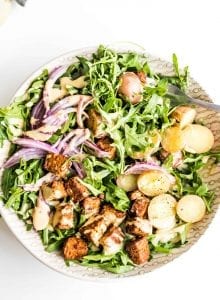 A bowl of arugula salad with potato, red onion and tempeh.