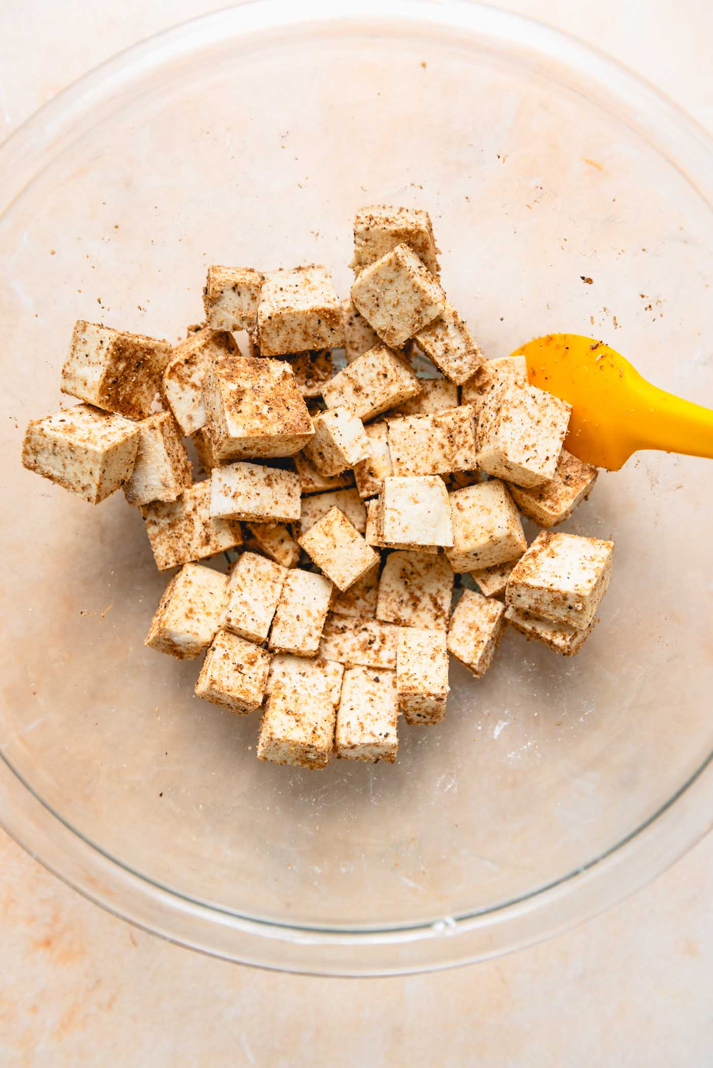Tofu cubes mixed with spices and cornstarch in a clear glass mixing bowl, ready for cooking.