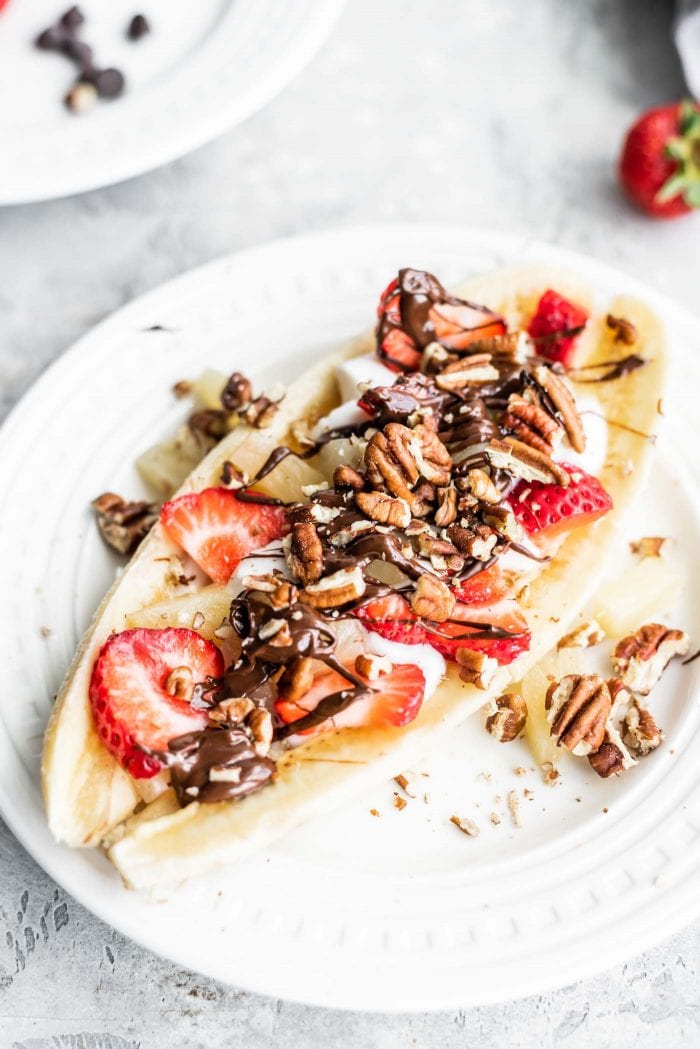 A banana sliced in half lengthwise topped with strawberries, pecans, yogurt and a drizzle of chocolate on a plate.