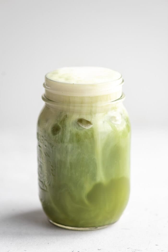 Close up of an iced matcha latte in a glass jar against a grey background.