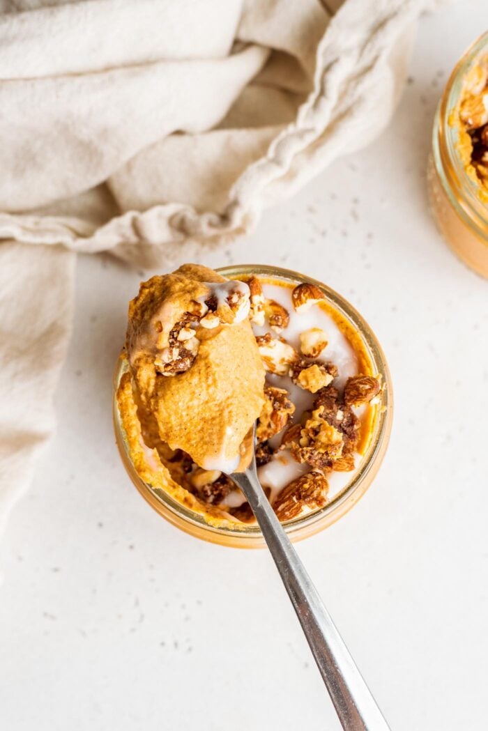 Spoon scooping pumpkin mousse and pecan crumble out of a small, glass jar sitting on a white surface with a dish cloth in the background.
