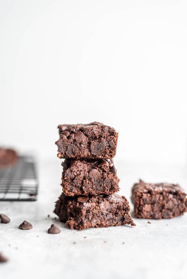 Stack of 3 vegan oat flour brownies on a grey surface with some chocolate chips scattered around.