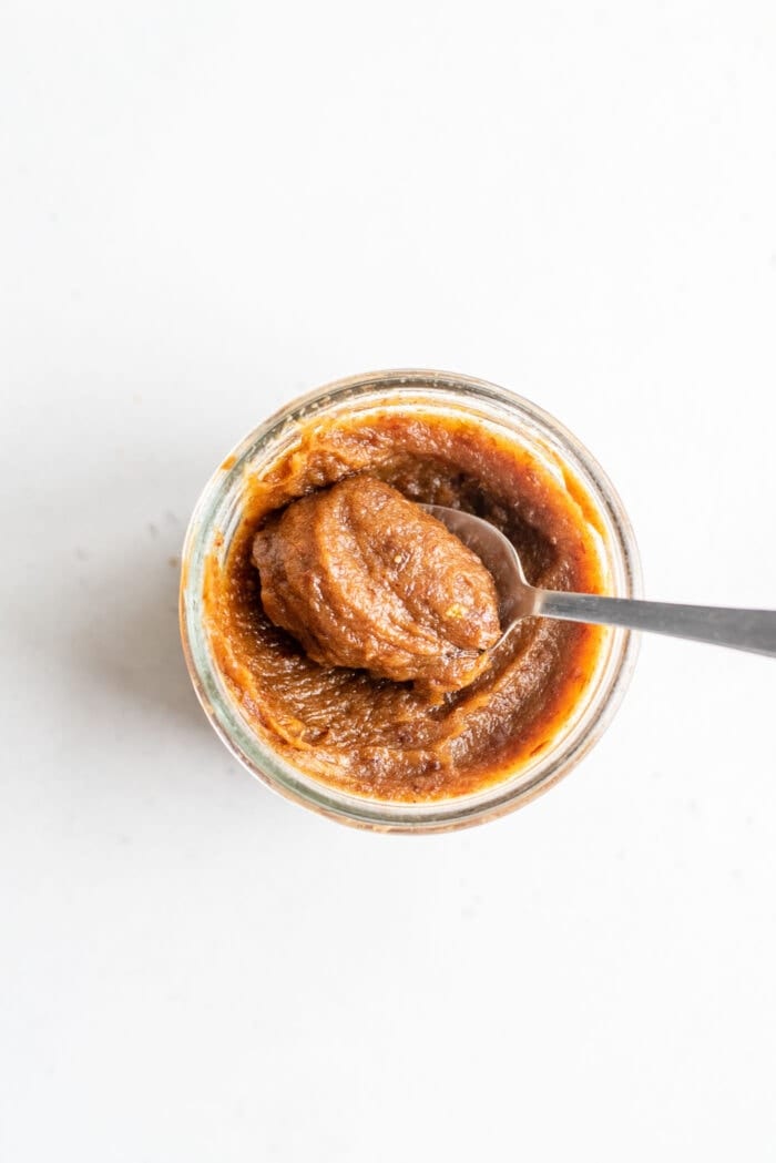 A spoon scooping date paste out of a small glass jar on a white background.