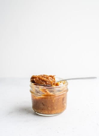 A spoon scooping date paste out of a small glass jar on a white background.