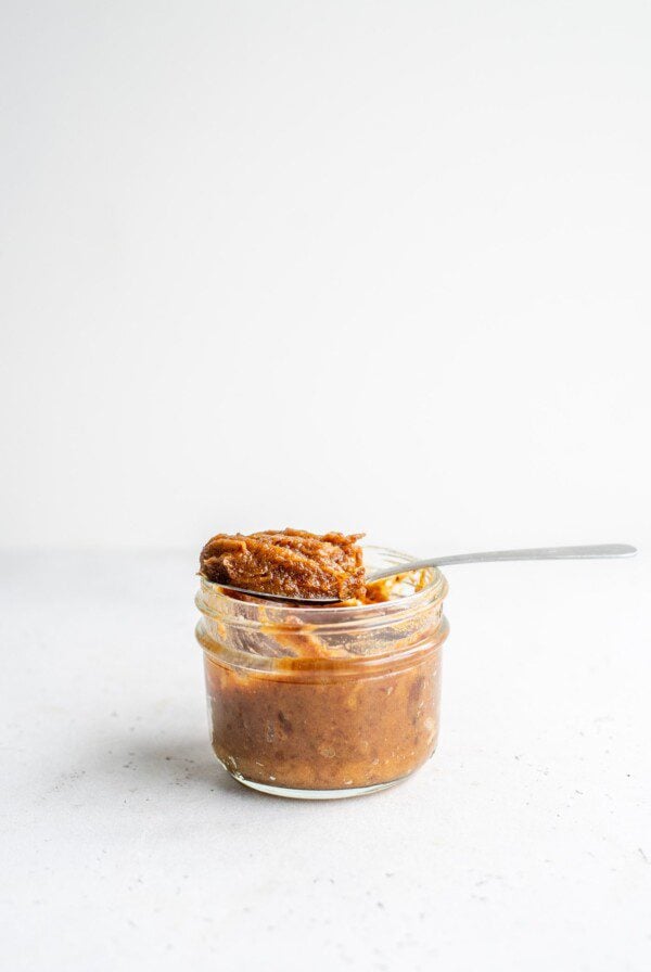 A spoon scooping date paste out of a small glass jar on a white background.