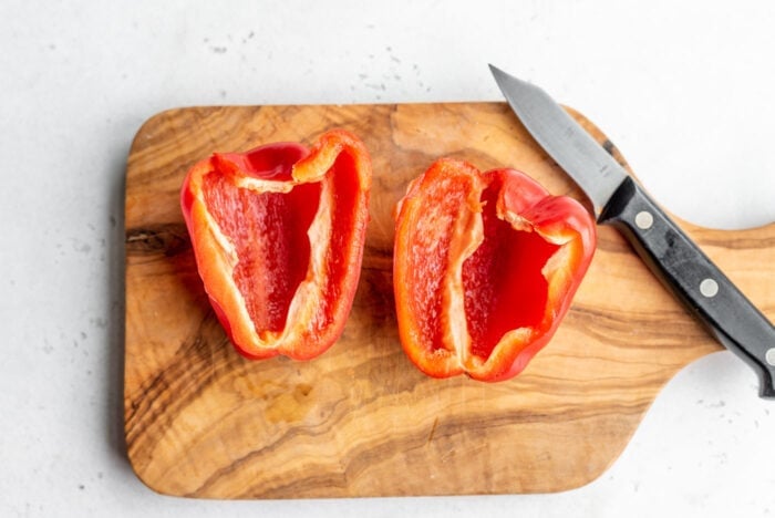 A red pepper cut in half with the seeds removed on a cutting board with a knife.