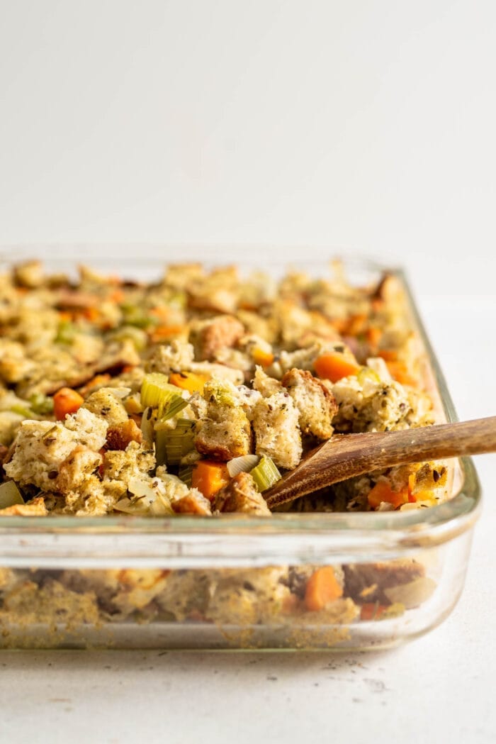 A wooden spoon scooping bread and vegetable stuffing out of a glass casserole dish.
