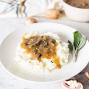 Mushroom gravy over mashed potatoes on a plate with some fresh sage on it.