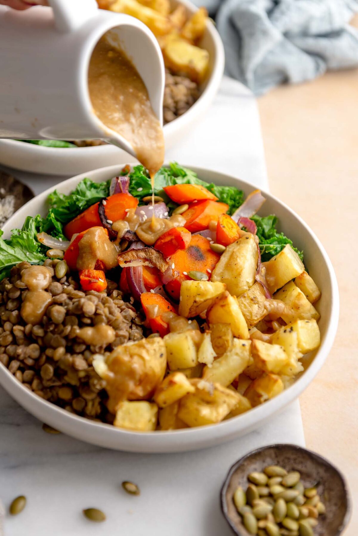 Pouring gravy from a gravy boat over a bowl with lentils and chopped, roasted potato, carrot, onion and kale.