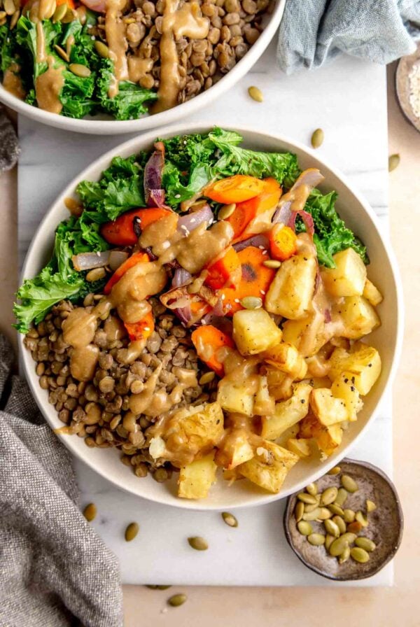 Bowl with roasted potatoes, carrots and onion, kale, lentils and a creamy sauce on top. It's sitting on a cutting board with a small dish of pumpkin seeds beside it.