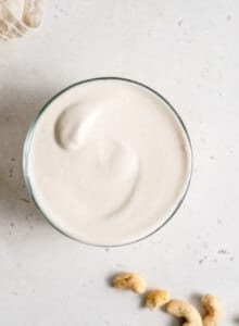 Overhead shot of thick cashew cream in a small glass dish with some raw cashews sitting beside it.
