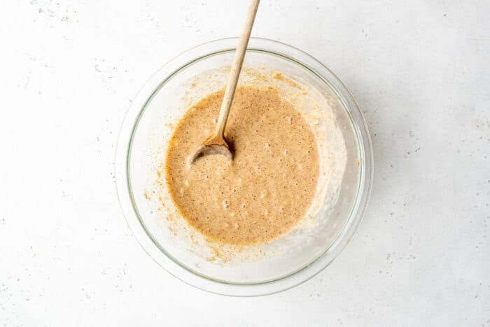 Mashed banana, almond butter and plant-based milk in a glass mixing jar with a wooden spoon.