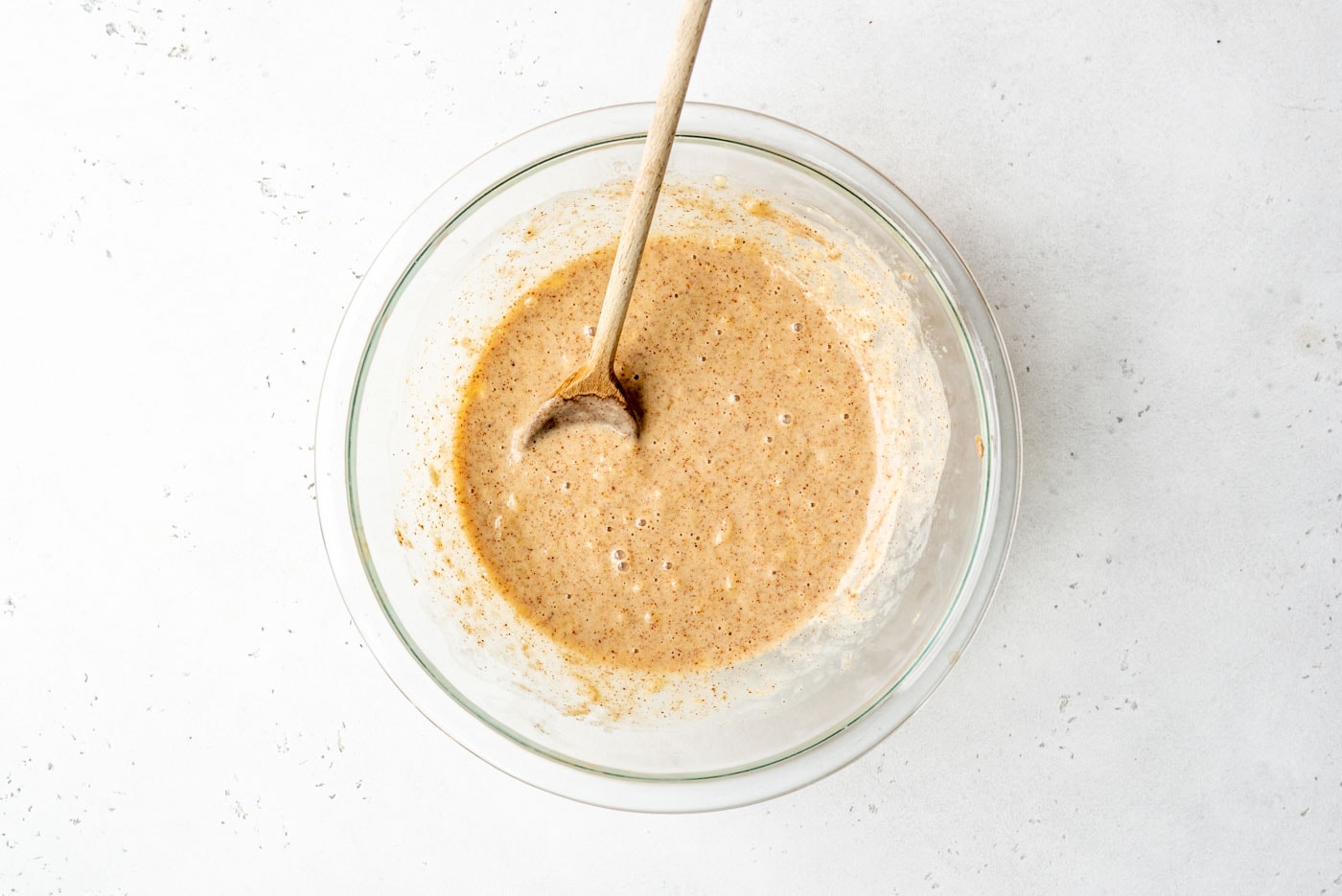 Mashed banana, creamy almond butter, and wholesome plant-based milk gently stirred in a glass mixing bowl with a wooden spoon, forming the wet base for the oatmeal cups.