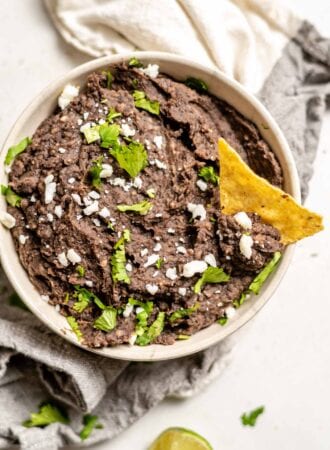 A bowl of vegan refried black beans with a chip in it topped with cheese and cilantro.