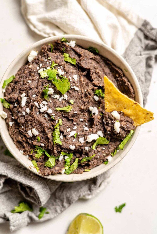 A bowl of vegan refried black beans with a chip in it topped with cheese and cilantro.