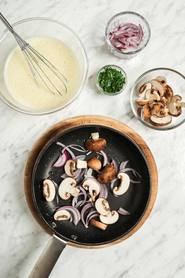 Mushrooms and onions cooking in a hot pan. A glass mixing bowl of batter sits beside the pan.