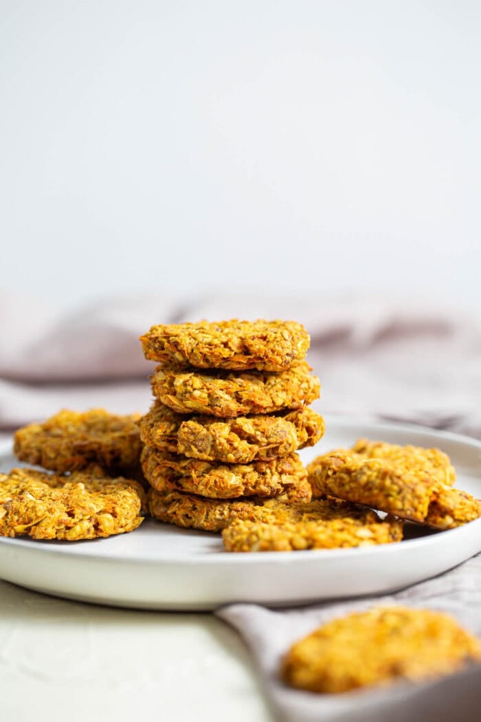 Five turmeric carrot oatmeal cookies stacked on a plate with other turmeric cookies around it.